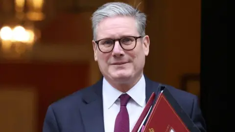 Sir Keir Starmer.  Seen from the shoulders up, her is wearing glasses and a navy suit with purple tie and carrying a red folder, looking straight ahead as he walks out of Downing Street 