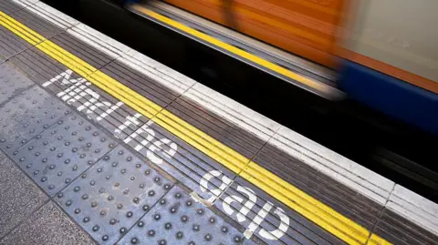 Close-up view of a Tube station platform featuring a yellow line and the words "Mind the gap" printed in white letters. The bottom of some train doors can also be seen.