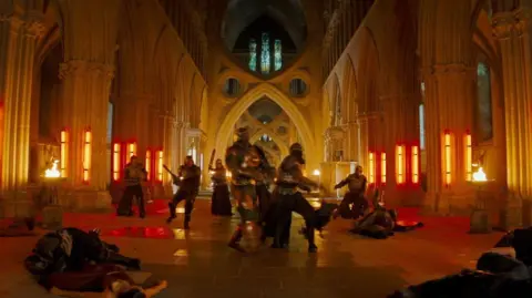 Amazon MGM Studios A still from Masters of the Universe. There is a group of actors in superhero costumes stood inside Wells Cathedral. The building is tall with arches and stained glass windows. 