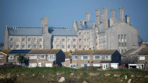 A general view of HMP Portland - a large grey building that bears over a row of houses. 