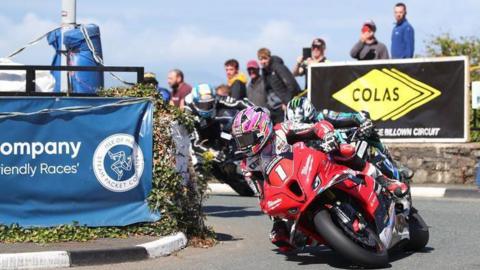 Davey Todd racing on a red motorcycle wearing red and white leathers and a pink helmet. Michael Dunlop is behind him on the road wearing black, purple and turquoise leathers and helmet. There are sponsors' banners in the background and spectators watching from behind a stone wall. Todd's bike had a red plate with the number one in white on the front.