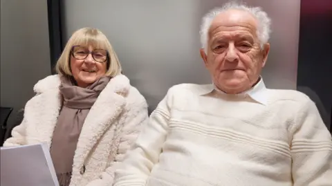 The couple are sat together smiling at the camera. John has a cream jumper on and a white shirt and has grey hair. His wife has a brown top on with a white fluffy coat over the top. She is also wearing glasses.