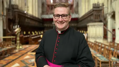 A member of the clergy stands inside Gloucester Cathedral, wearing traditional black robes with red trim, with the ornate choir stalls and vaulted ceiling visible in the background.