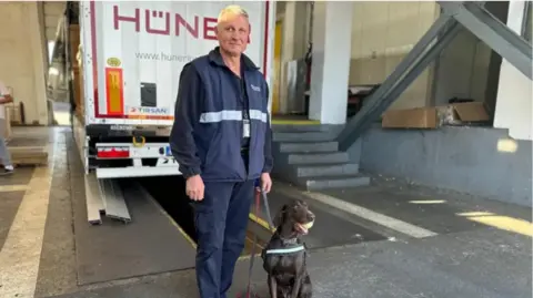 Adele the sniffer dog is standing with her handler beside a lorry in a customs shed on the Bulgaria - Turkey border.