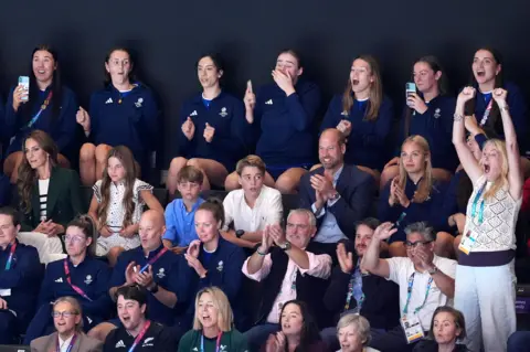 PA Media The Duke and Duchess of Rothesay with their three children watching a netball match. Everyone is seated while cheering, apart from a blonde woman on the far right of the photo who is standing with her arms in the air. She appears to be cheering loudly.
