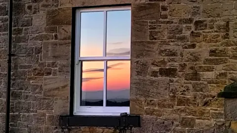 BBC Weather Watchers/Wellibobs A white window on a stone building with the reflection of the red and orange sunrise or sunset on it. Wispy clouds are also reflected.