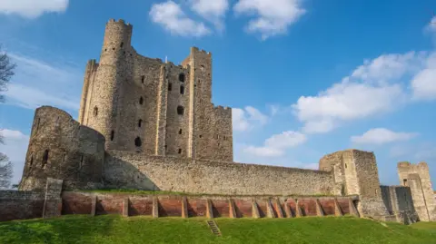 An ancient castle with an area of green grass in front of it and blue sky above it. 