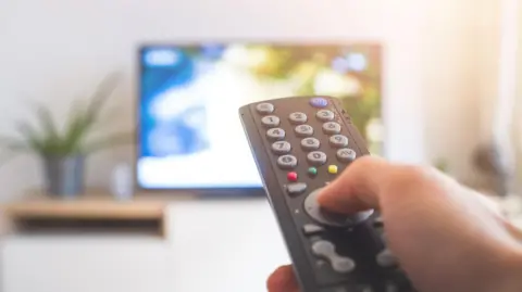 Getty Images A stock image of someone watching TV. You can see a hand holding a black remote control, the background is out of focus but you can see the outline of a TV screen.