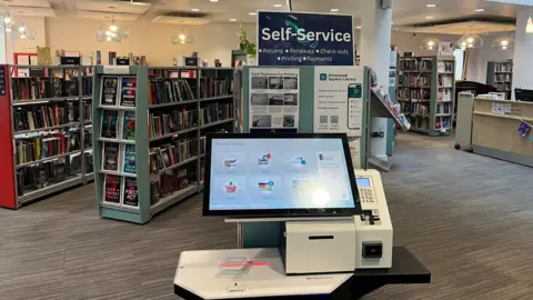 A library checking books in and out machine has a large "self service" sign above it in amongst rows of bookshelves. The room is well lit with a reception desk to the right.