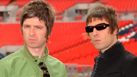 PA Media Liam and Noel Gallagher in 2008. Noel has brown hair and is wearing a green jacket. Liam also has brown hair, is wearing dark sunglasses, a black polka dot shirt and a black jacket. They appear to be standing in a football stadium, with seats behind them.