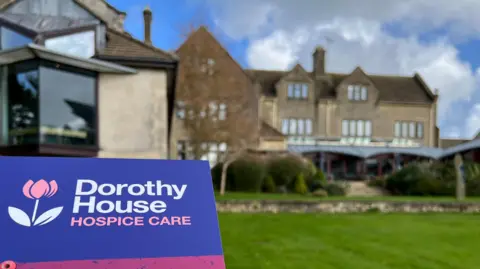 Dorothy House's main base at Winsley, near Bradford-on-Avon. It is a large house can been seen with lush green garden under a blue sky, but the focus is on a Dorothy House Hospice Care sign.