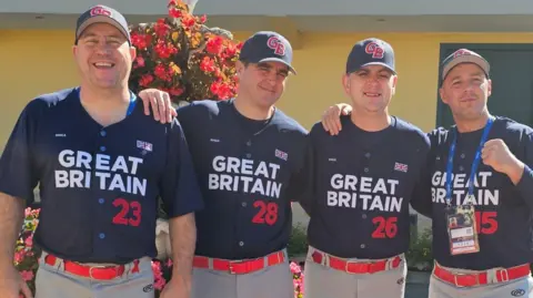Ray Clements Ray smiles whil standing alongside three other men in the team. They all wear dark baseball caps and uniform shirts with Great Britain written across them. They pose in front of a flower display and building at a tournament.