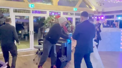 A man in a white beanie hat congratulates a newly married couple on a dance floor