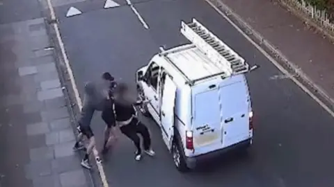 Bedfordshire Police An ariel CCTV view of Havelock Road in Luton. A small white van with a ladder on the roof is stopped in the middle of the road, with a side door panel open. Next to the door, two men try to drag a third into the van. Their faces are all blurred out and you can see three pairs of feet. The victim wears black jogging bottoms with white stripes.