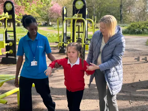 Vanellope is walking in the park with her mum holding one arm and a carer holding the other 