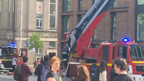A close up of firefighters at a scene outside a block of flats in Bristol. Two people can be seen walking down a fire engine ladder and onlookers stand in the street.