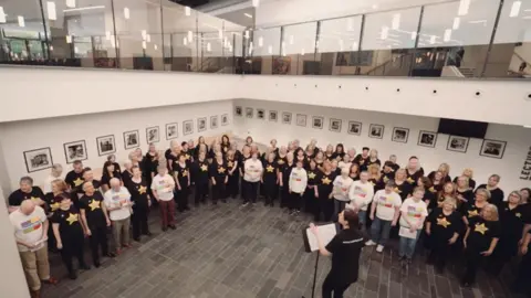 Dementia NI Members of the Dementia and Rock Choir in black and white t-shirts performing The Climb being led by choir leader, Rachel Coulter. 