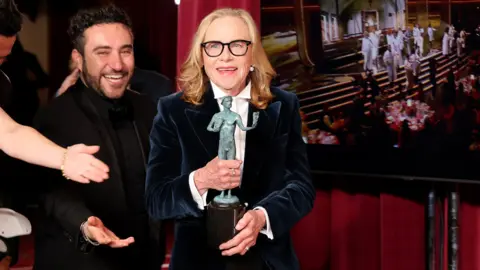 Amy Madigan backstage with her award at the 32nd Annual Actor Awards in Los Angeles. She is wearing a navy velvet jacket and white shirt and there are smiling people next to her and screen showing the ceremony to the right