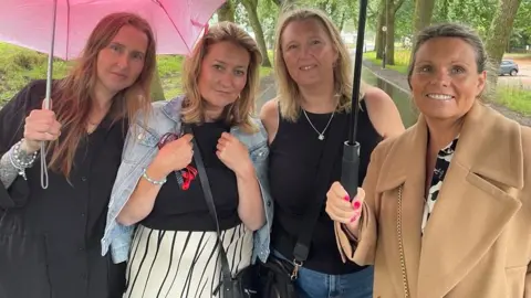 Cuatro mujeres se pararon en un camino en un parque. Uno sostiene un paraguas rosado y tiene un vestido negro, con el pelo largo y marrón claro, la segunda izquierda sostiene su chaqueta de mezclilla alrededor de sus hombros, sobre su top negro y tiene una falda a rayas en blanco y negro. A su derecha, la mujer tiene cabello rubio y un chaleco negro. Ella también usa un collar colgante de plata. A la derecha, la mujer sostiene un paraguas y lleva un abrigo de camello.