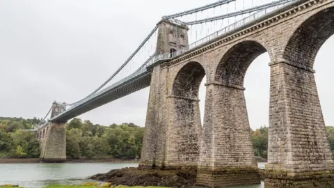 Menai Bridge which is made of bricks and is over a body of water. Trees can be seen on the opposite bridge. 