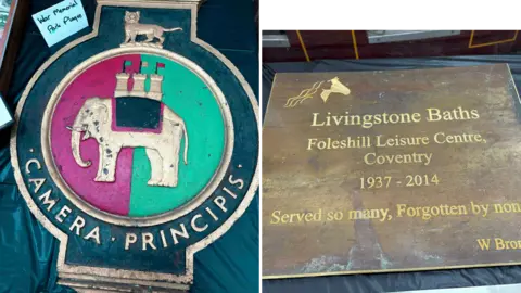 A War Memorial Park plaque shows an elephant crest in the middle of a bright pink and green circle. It has Camera Principis in gold lettering. To the right of the picture a bronze plaque reads: "Living stone Baths, Foleshill Leisure Centre, Coventry - 1937-2014. Served by so many, fotgotten by none