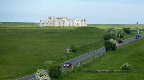 A303 with cars travelling along with Stonehenge in the distance. You can also see large fields on either side.