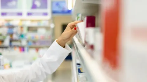 Woman takes a box of medicine from a shelf lined with boxes of medicine. She has red nail varnish on her nails.
