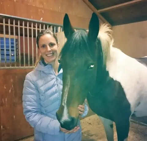 Kirstie Pickles Kirstie poses happily with a horse in a stable. She wears a thick winter coat, light blue in colour. The horse has a mainly brown head, with white parts on its shoulders and legs.