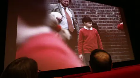 A cinema screen shows a young boy in a plain red school jumper standing in a playground next to a teacher in a grey jacket, red tie and white shirt. A man with thinning black hair and a woman with black hair and glasses watch on from a set of red cinema seats.