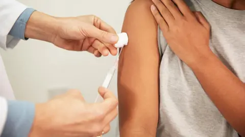A woman raises the arm of her grey T-shirt as a doctor injects a needle into her arm while holding cotton wool against it