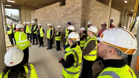 Around 30 people wearing white hard hats and hi viz vests are standing in a building site.