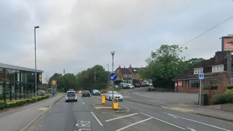 Road junction with crossing island and traffic coming bothways. A pub is to the right and Lidl store on the left. The road has a crossing island in the centre.
