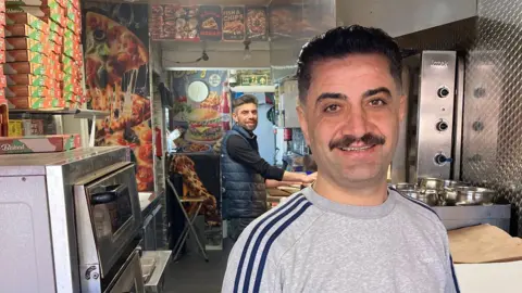 Sean Dunderdale/BBC A man is standing behind the counter in a takeaway food shop, smiling at the camera. He has short black hair and a moustache. He is wearing a grey T-shirt with three blue stripes on the arms. Behind him is another man, also smiling at the camera. He is wearing a long-sleeved black T-shirt, a blue padded bodywarmer and black trousers. He has short black hair. There are lots of cooking appliances, pizza boxes and pictures of food on the walls.