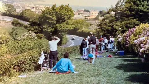Glen Kinrade And old photograph of crowds watching from a garden over a hedge. A motorbike is coming round a corner.