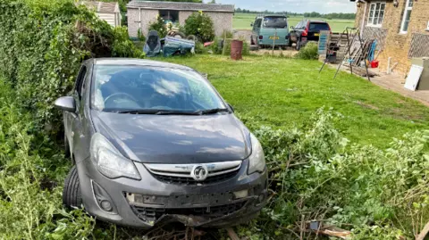 A grey Vauxhall Astra crashed into the garden - with damaged front - the number plate has fallen off- and plants and a hedge have been damaged 
