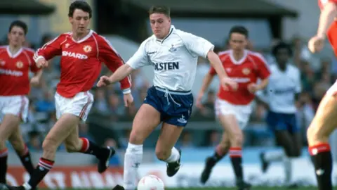 Getty Images Paul Gascoigne is wearing a white Tottenham Hotspur shirt, blue shorts and white socks. Across the front of the shirt is the logo "Holsten". He is being pursued by Manchester United players wearing red shirts featuring the "Sharp" logo.