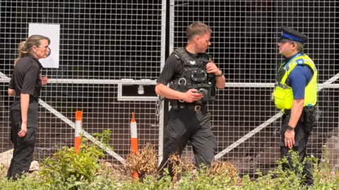 BBC Police stand near silver gates with silver wire above the gates. A woman is in black on the left and two men, including one in a yellow hi-vis jacket, are on the right of the photo.