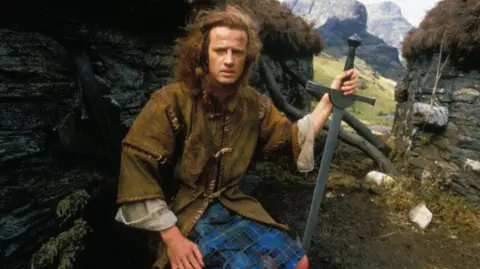 Georges De Keerle/Gamma-Rapho via Getty Images Christopher Lambert is wearing period Highland garb and is holding a sword as sits by the wall of a stone walled, turf roofed building. A glen and mountains are behind him.