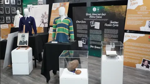 Several glass cases with rugby related memorabilia including a book, a photo and two rugby balls are on white plinths. Behind them is a mannequin wearing a green, navy and yellow rugby top. There are also lots of information boards in the background. 