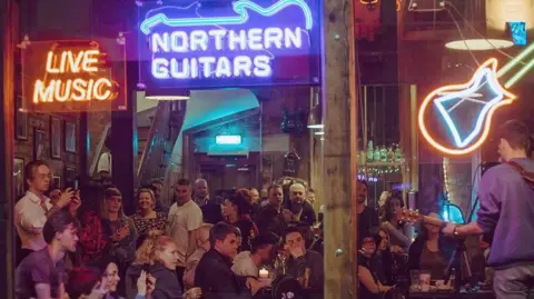 The outside of Northern Guitars music venue in Leeds is pictured. There are three neon signs in the window - one saying 'live music', one saying 'Northern Guitars' and one with the silhouette of a guitar. A man is playing the guitar and singing, as a crowd looks on.