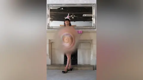 Deafblind UK A woman is standing in front of a front of a fireplace and a mirror smiling, wearing a pink hat and is naked.