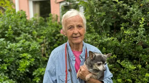 Maggie Jackaman, who has short grey hair and is wearing a light blue and white striped shirt over a pink top, holding her dog, Kenny. She is standing in front of a hedge.
