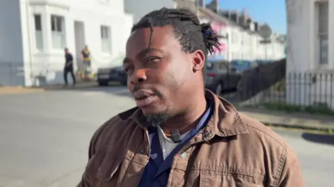 A man stood looking directly at the camera. He is wearing a brown jacket. 