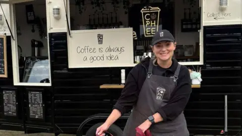 A woman wearing an apron and baseball cap poses for a photo at a country show outside her coffee trailer. She is holding onto two sheep who are wearing headcollars and leads.