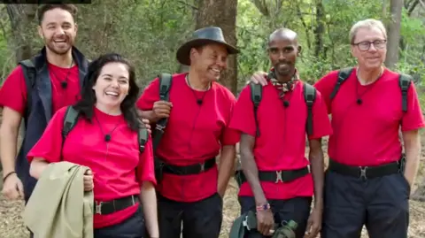 Adam Thomas, Scarlett Moffatt, Craig Charles, Mo Farah and Harry Redknapp standing together, smiling and all wearing red I'm A Celebrity T-shirts