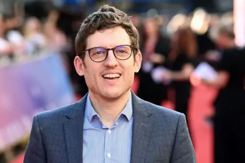 Getty Images Elis James has brown hair, black glasses and is smiling. He is wearing a blue blazer and blue, striped shirt