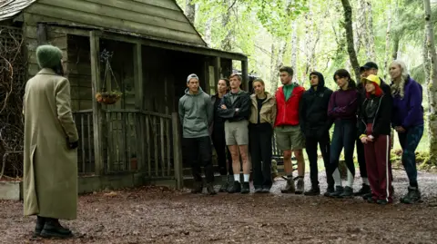 BBC/Studio Lambert/Euan Cherry A picture of the cast of the Traitors stood outside a wooden cabin.