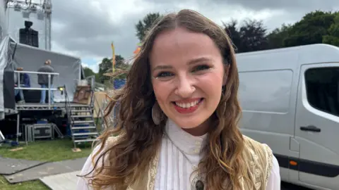 BBC Wales Mari smiles broadly to camera. She is backstage at a festival and wears her hair loose around her shoulders. She has a nose piercing and wears oversized metal earrings. It is a head and shoulders shot and she has a white top on.