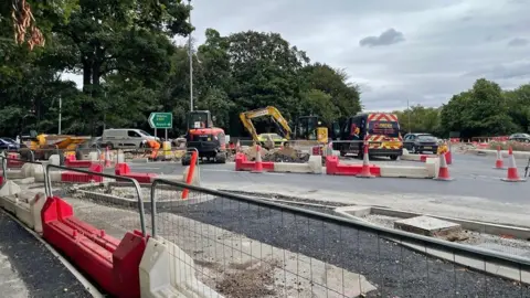 Roadworks at a large road junction, with excavators, construction workers in high-visibility clothing, cones and red-and-white barriers surrounding excavated sections of roadway. Newly laid tarmac and unfinished kerbs are visible in the foreground, while cars, a contractor van and a green motorway direction sign stand against a backdrop of trees under a cloudy sky.