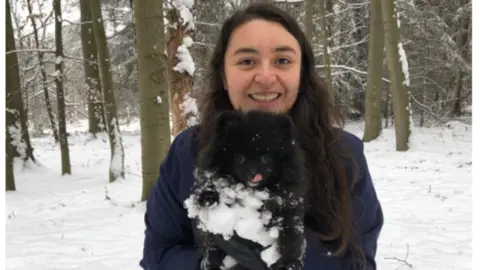 family handout Maria holds a little black fluffy dog and smiles at the camera. They are standing in woods that are covered in snow, and the dog has snow on its tummy. Maria wears a blue fleece and has very long black wavy hair.